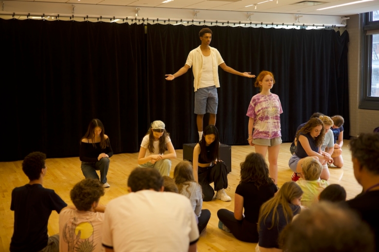 A group of Tisch Summer HIgh School Drama students pose on a stage during their final presentations on the last day of class. One student stands on a raised box with arms out to his side while other students kneel or stand beside and in front of him